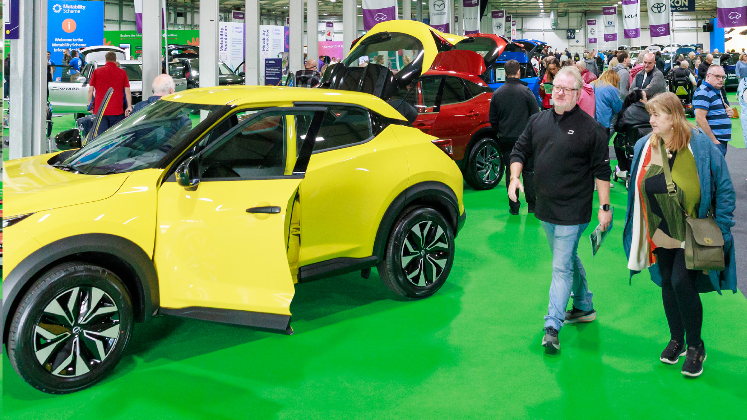 Visitors walk through a busy Motability Scheme exhibition hall, passing a bright yellow car with its door and boot open for viewing.