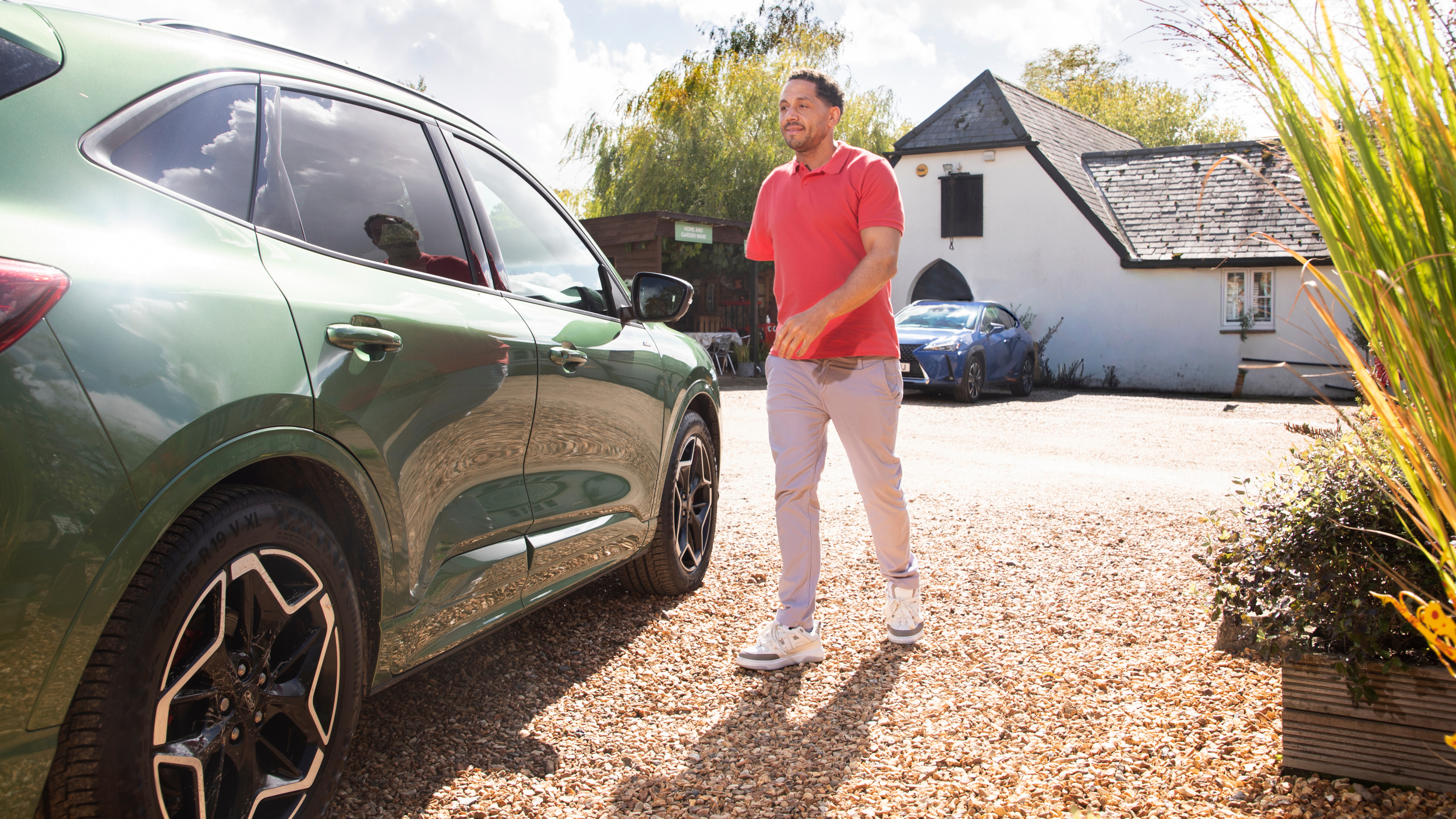 A man walks across a gravel driveway towards a green electric SUV parked outside a small white building, with trees and another car in the background on a sunny day.