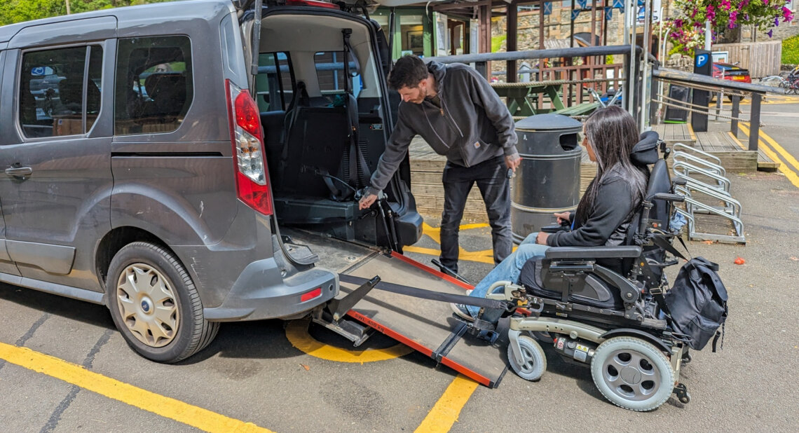 A man lowers a ramp from the rear of a grey Wheelchair Accessible Vehicle (WAV) while a woman using a powered wheelchair waits at the bottom of the ramp in a car park with buildings and bike racks in the background.