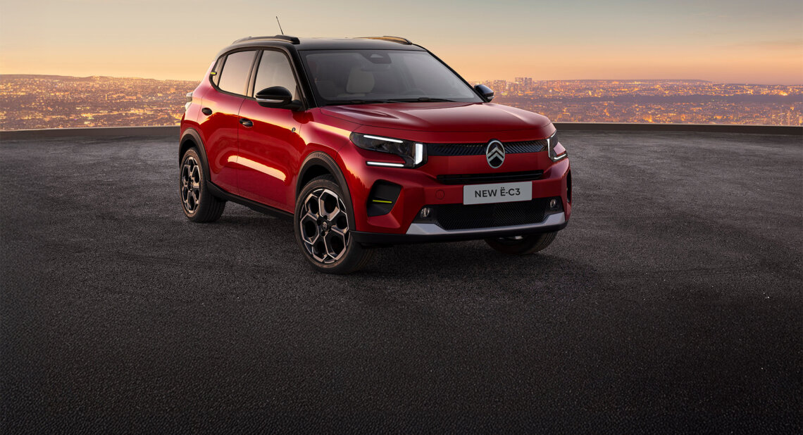 Citroën New ë C3 red electric compact SUV parked on an elevated surface at sunset with a city skyline in the background.