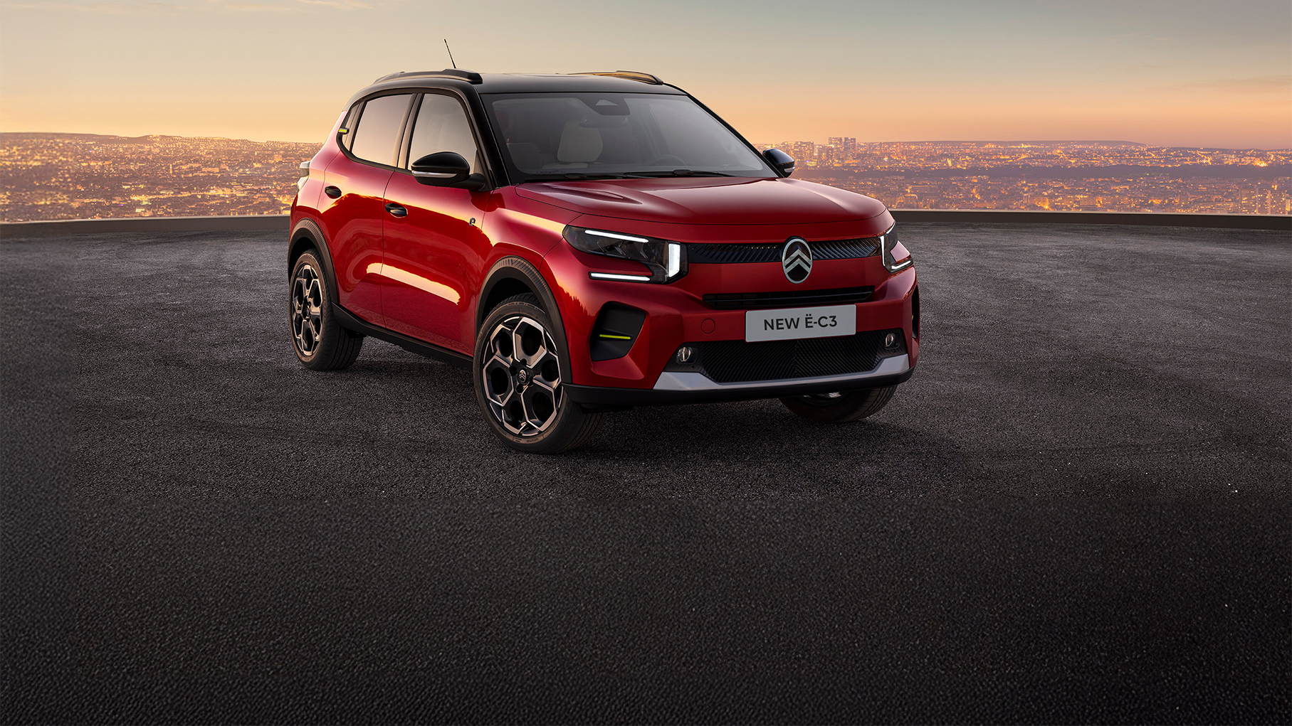 Citroën New ë C3 red electric compact SUV parked on an elevated surface at sunset with a city skyline in the background.