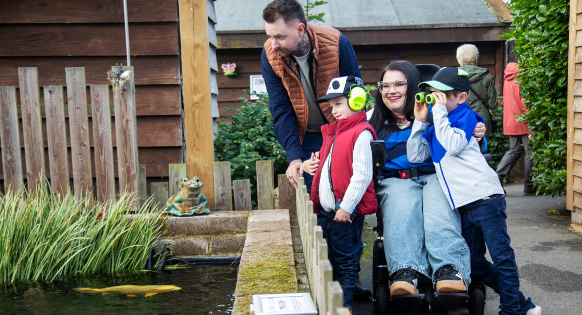 A family stands beside a garden pond, with a woman using a powered wheelchair smiling as two young children look into the water, one using binoculars and the other wearing ear defenders, while an adult leans in to join them.