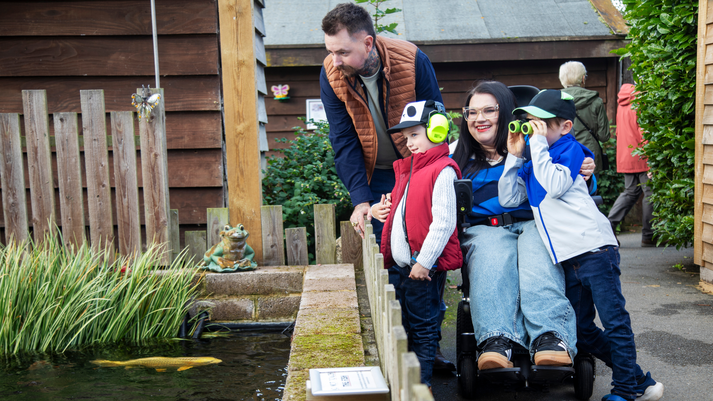 A family stands beside a garden pond, with a woman using a powered wheelchair smiling as two young children look into the water, one using binoculars and the other wearing ear defenders, while an adult leans in to join them.