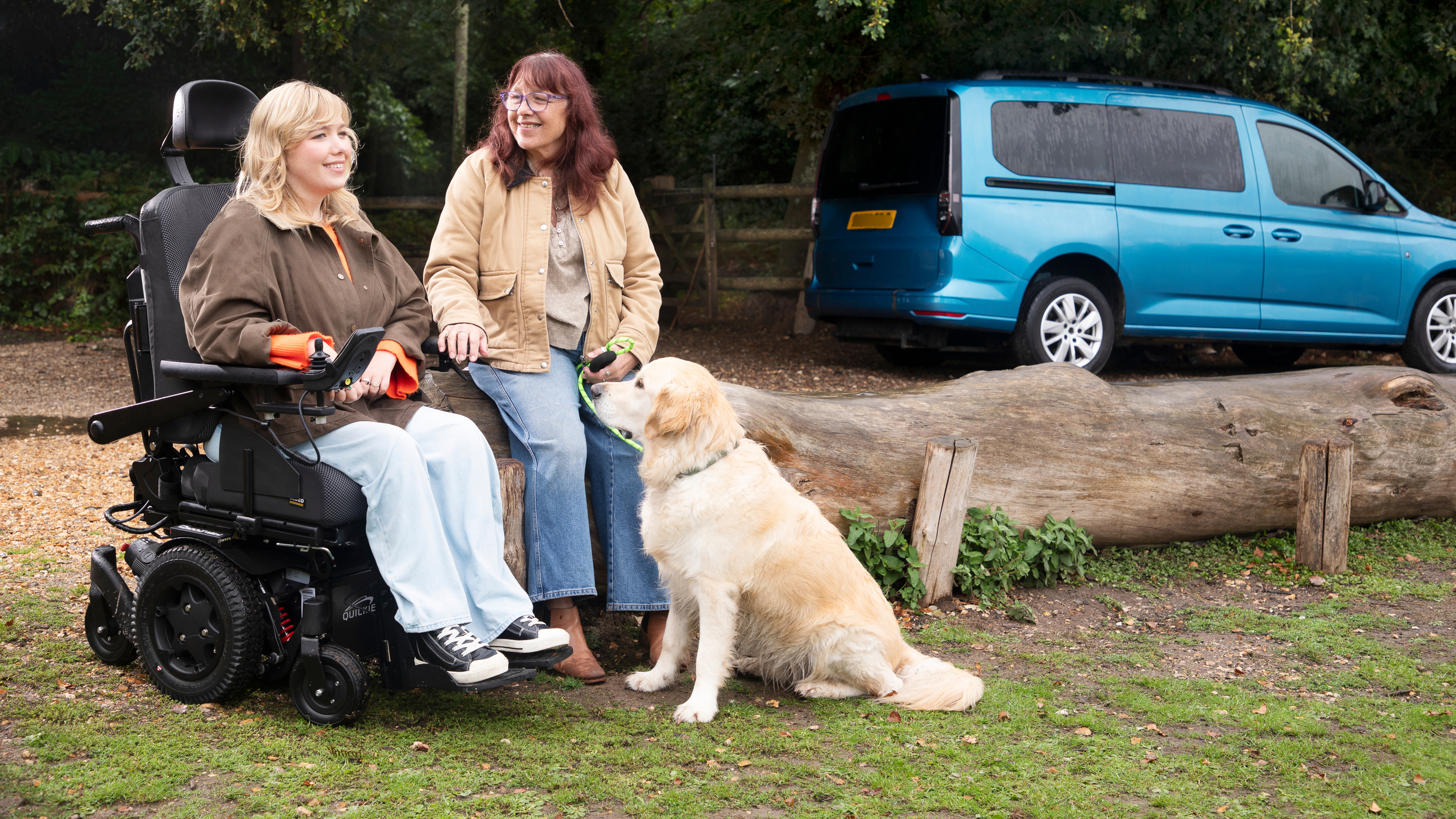 Two women pause on a woodland path, one seated in a powered wheelchair and the other standing beside her, while a golden retriever sits in front and a blue wheelchair accessible vehicle is parked behind them.