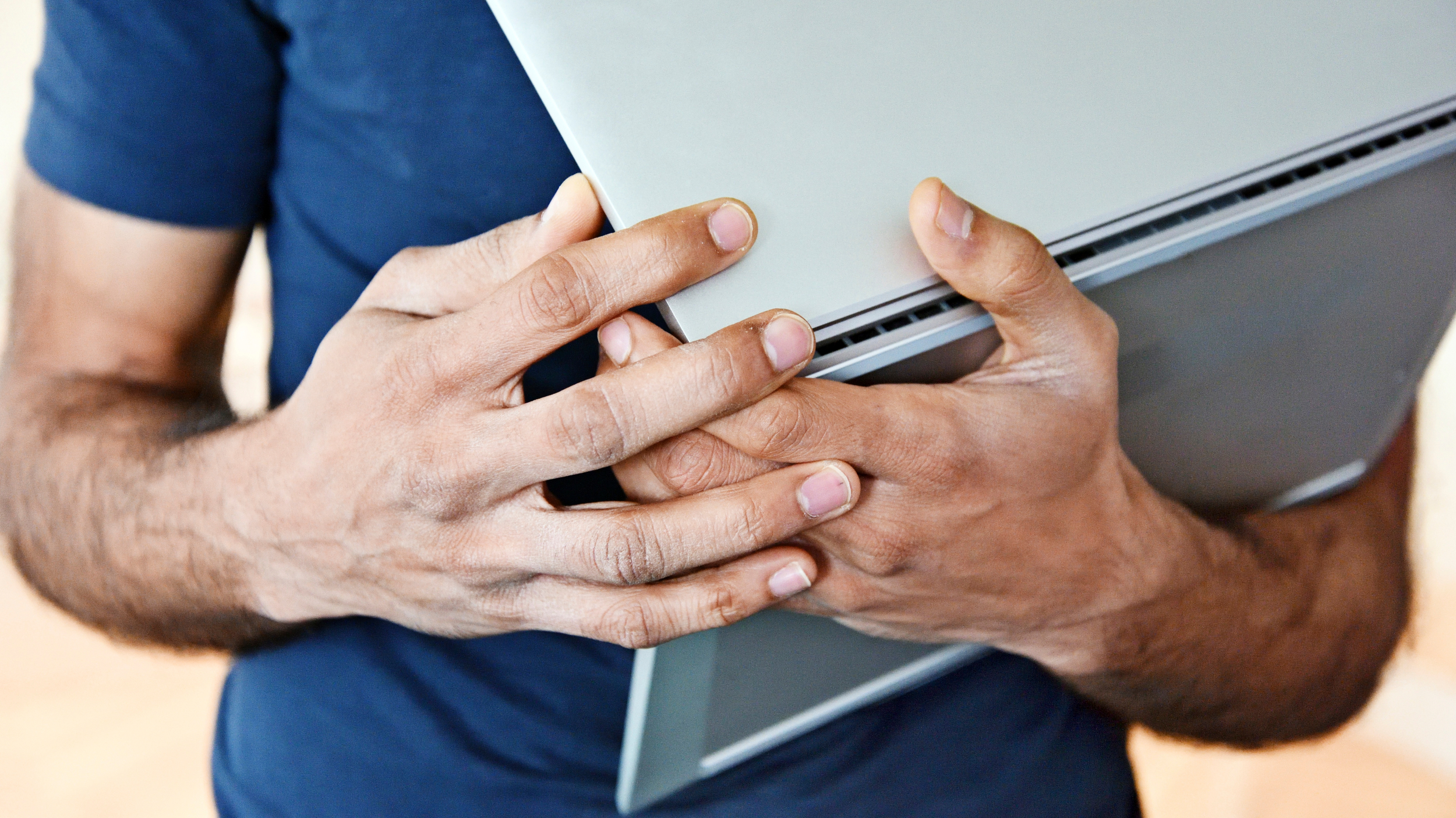 Close up of a person holding a closed silver laptop securely with both hands against their chest, wearing a blue short sleeved shirt.