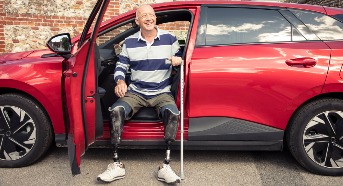 Smiling older man with double below knee prosthetic legs sitting in the driver’s seat of a red car with the door open, holding a walking stick, parked in front of a brick wall.