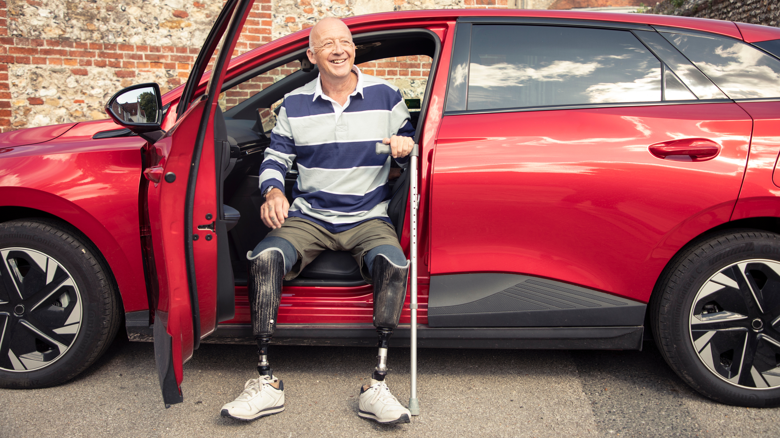 Smiling older man with double below knee prosthetic legs sitting in the driver’s seat of a red car with the door open, holding a walking stick, parked in front of a brick wall.