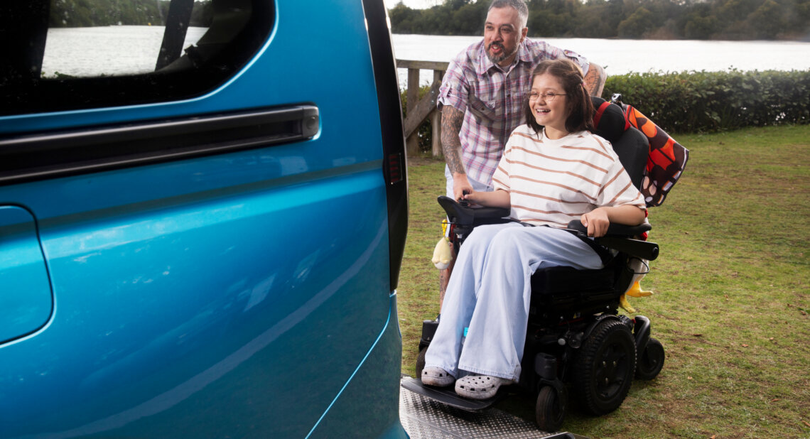 A smiling girl using a powered wheelchair is positioned on a lowered ramp at the back of a blue wheelchair accessible vehicle, with a man standing behind her. They are parked beside a lake with trees in the background.