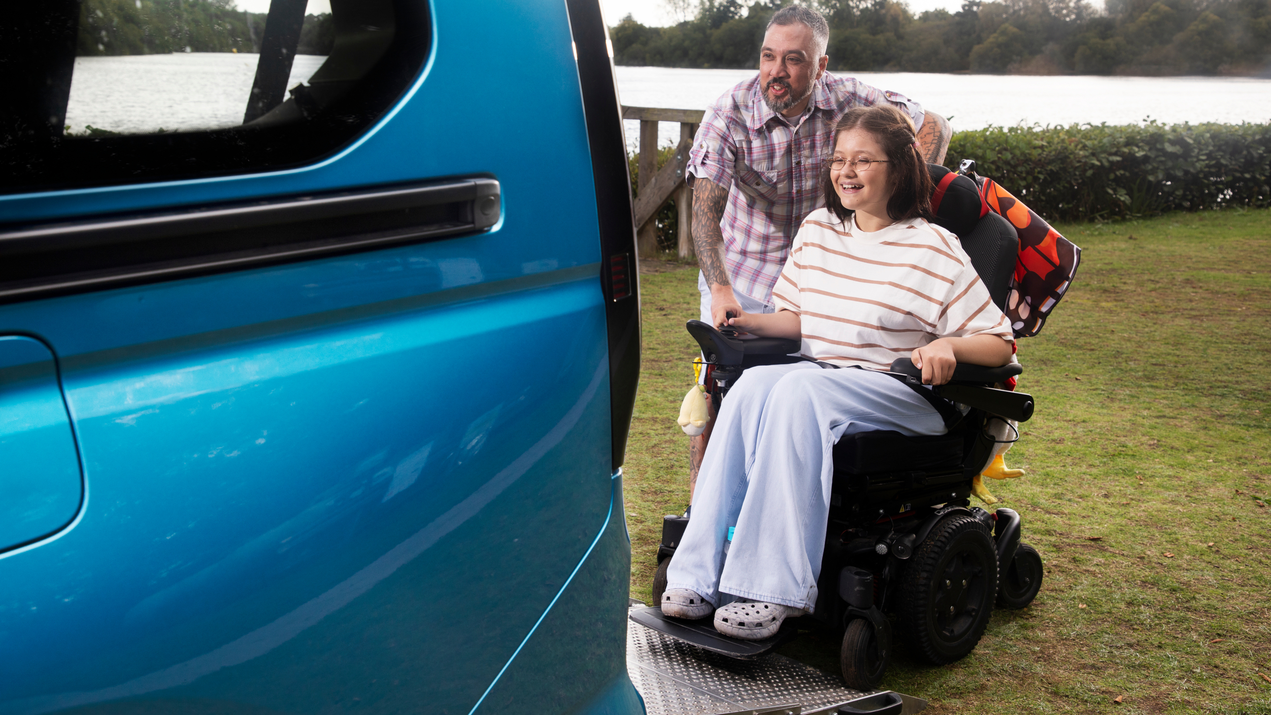 A smiling girl using a powered wheelchair is positioned on a lowered ramp at the back of a blue wheelchair accessible vehicle, with a man standing behind her. They are parked beside a lake with trees in the background.