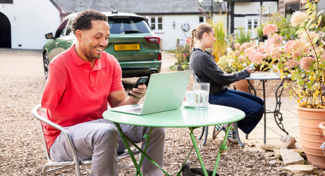 A man in a red polo shirt sits at a small green outdoor table, smiling at his phone with a laptop open in front of him and a glass of water beside it. Behind him, a green SUV is parked in a driveway outside a house. In the background, a woman sits at another table near pink flowering plants, also looking at her phone.