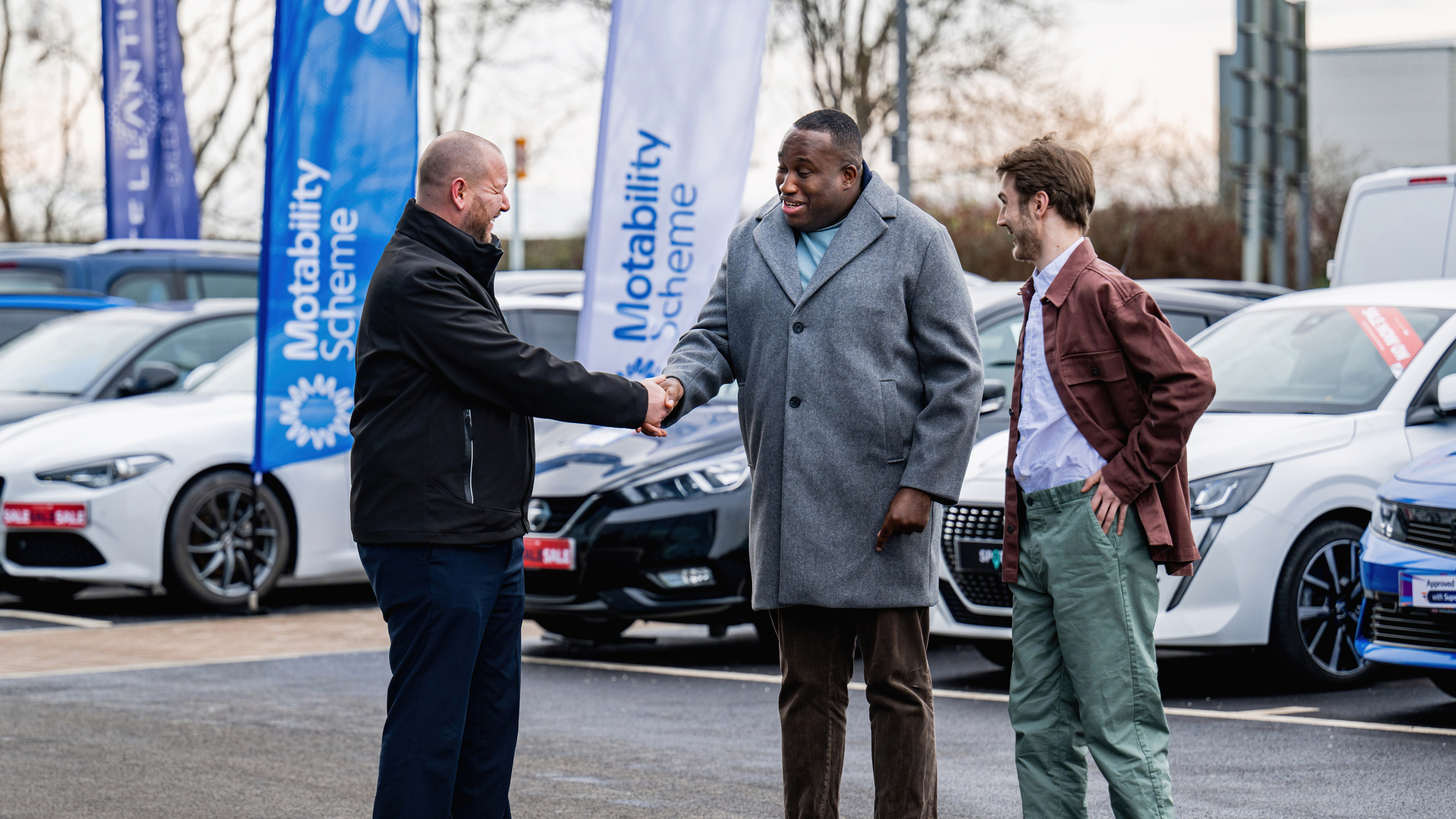 Three men standing in a car dealership forecourt shake hands in front of a row of parked cars, with blue Motability Scheme flags displayed behind them. One man in a black jacket greets a customer wearing a grey coat, while another man in a brown jacket stands beside them smiling.