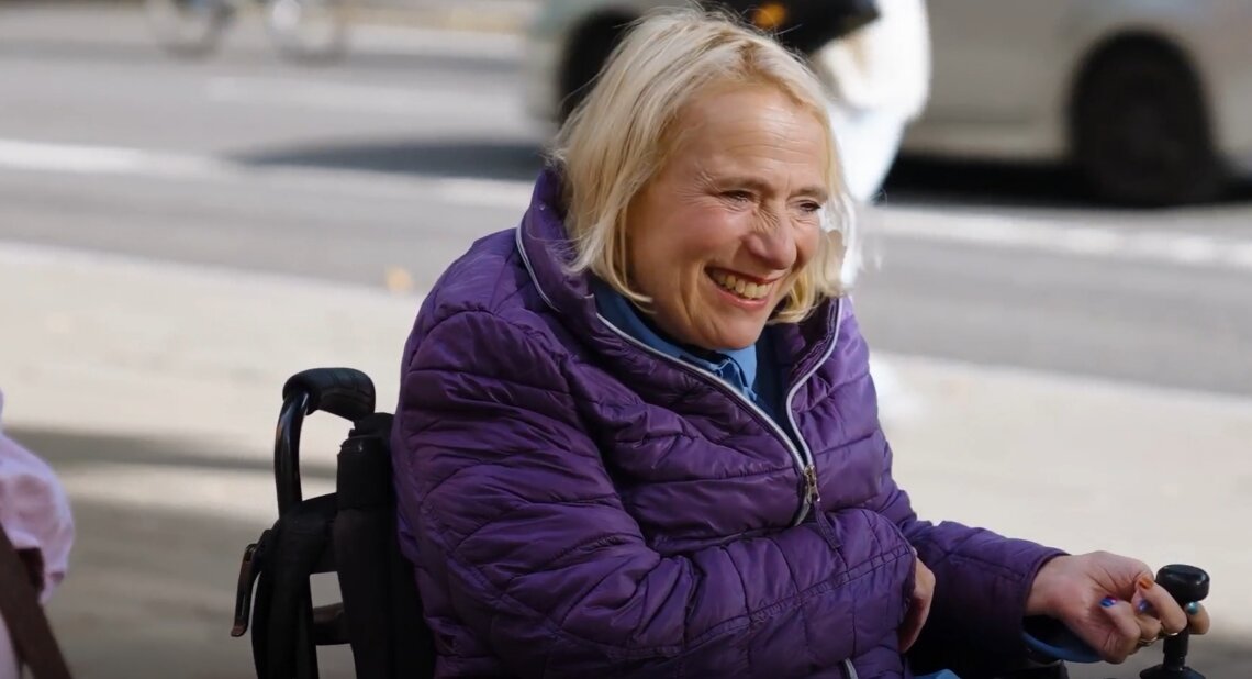 Alex Cowan, a Motability Scheme customer, smiling while using a powered wheelchair outdoors, wearing a purple jacket on a sunny street.
