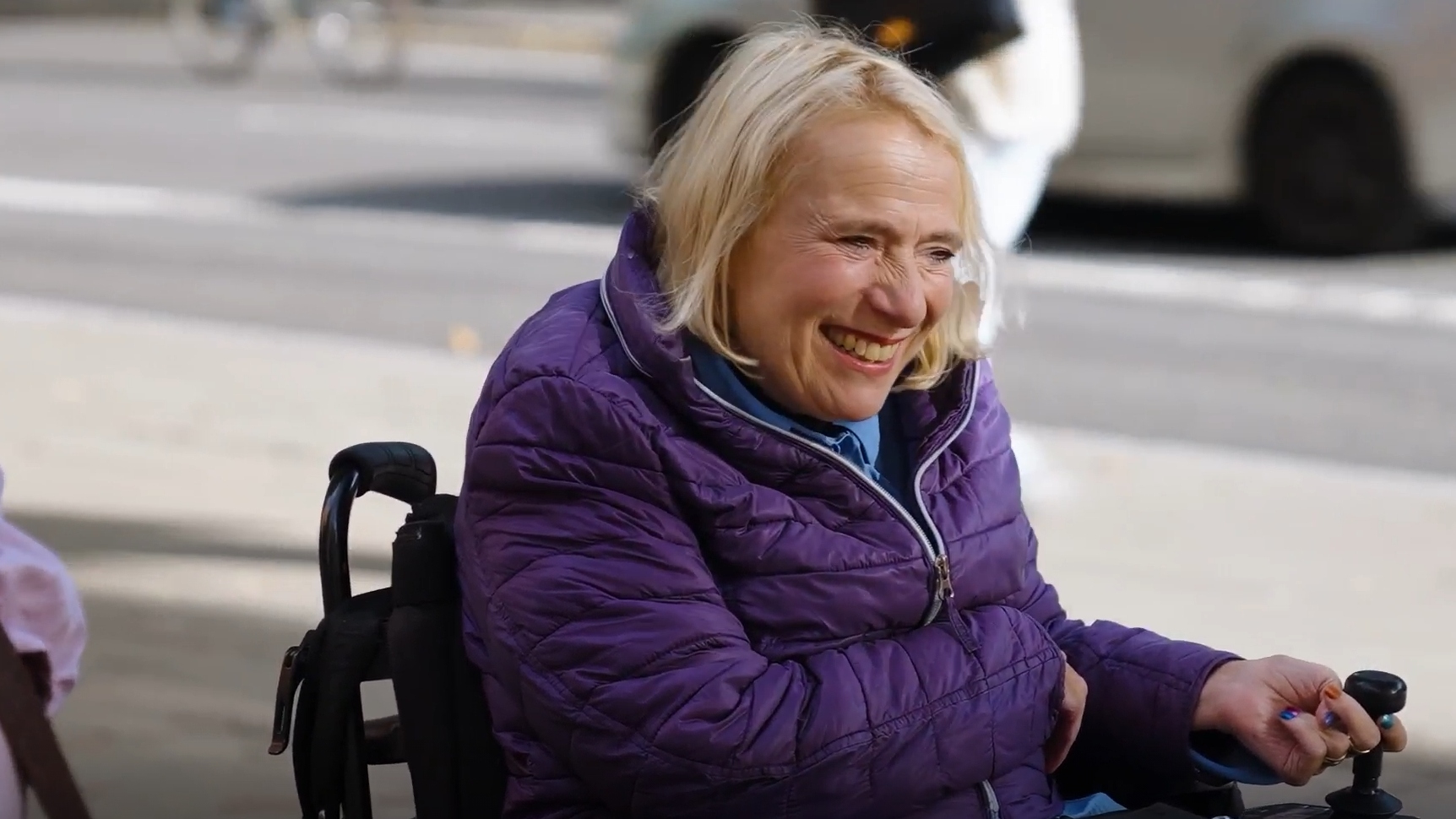 Alex Cowan, a Motability Scheme customer, smiling while using a powered wheelchair outdoors, wearing a purple jacket on a sunny street.