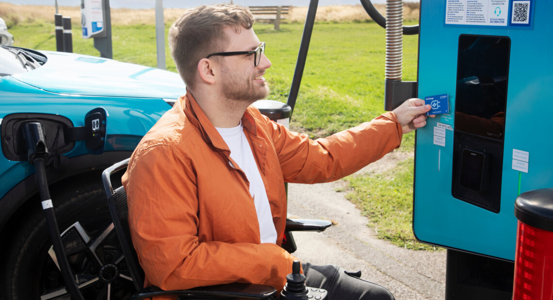 A man sitting in a powered wheelchair charges a turquoise electric car at a public charging point. He is smiling and holding a blue contactless payment card up to the reader on the charger. The car is plugged in beside him, and the scene is set outdoors near grass and a bench on a sunny day.