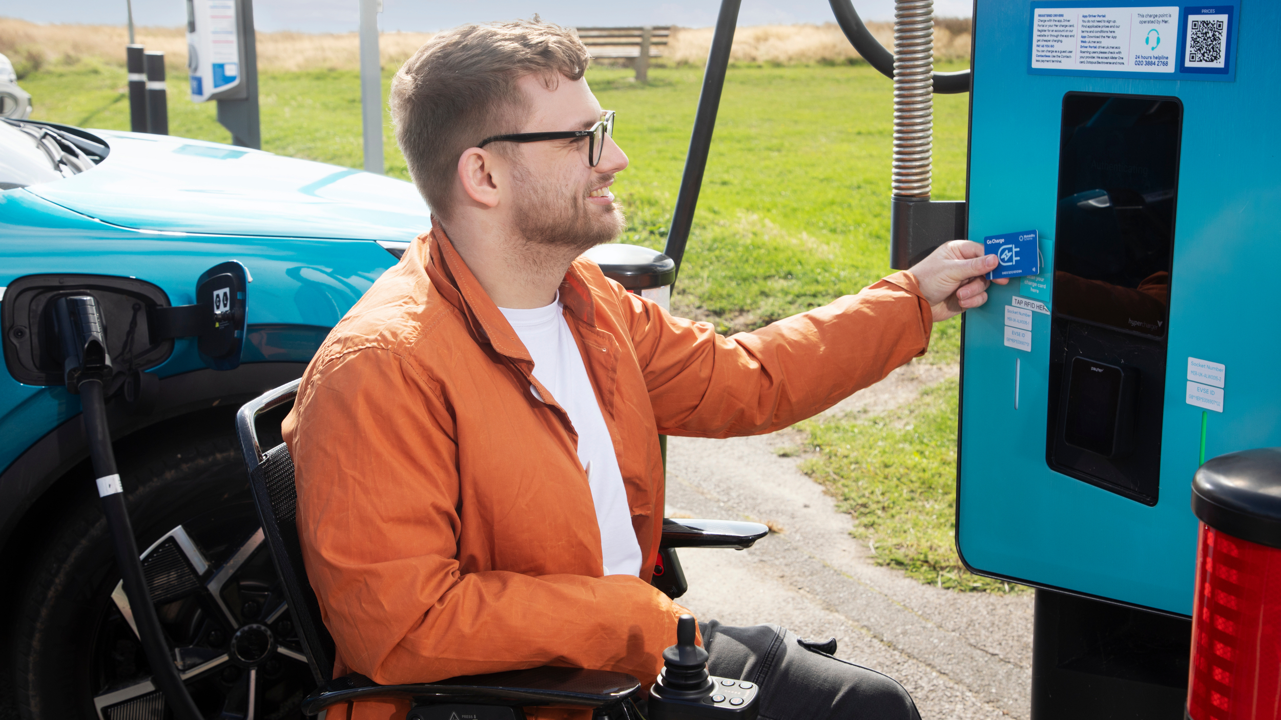 A man sitting in a powered wheelchair charges a turquoise electric car at a public charging point. He is smiling and holding a blue contactless payment card up to the reader on the charger. The car is plugged in beside him, and the scene is set outdoors near grass and a bench on a sunny day.