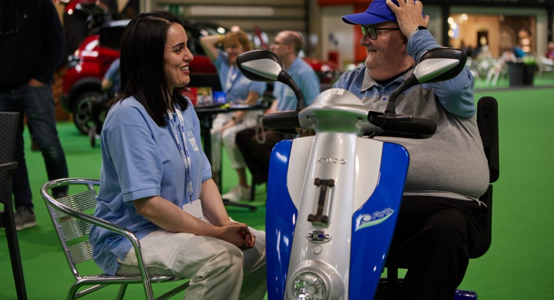 A man sits on a mobility scooter and smiles while talking to a staff member at Motability Scheme Live in Birmingham. Other visitors and vehicles are around them.