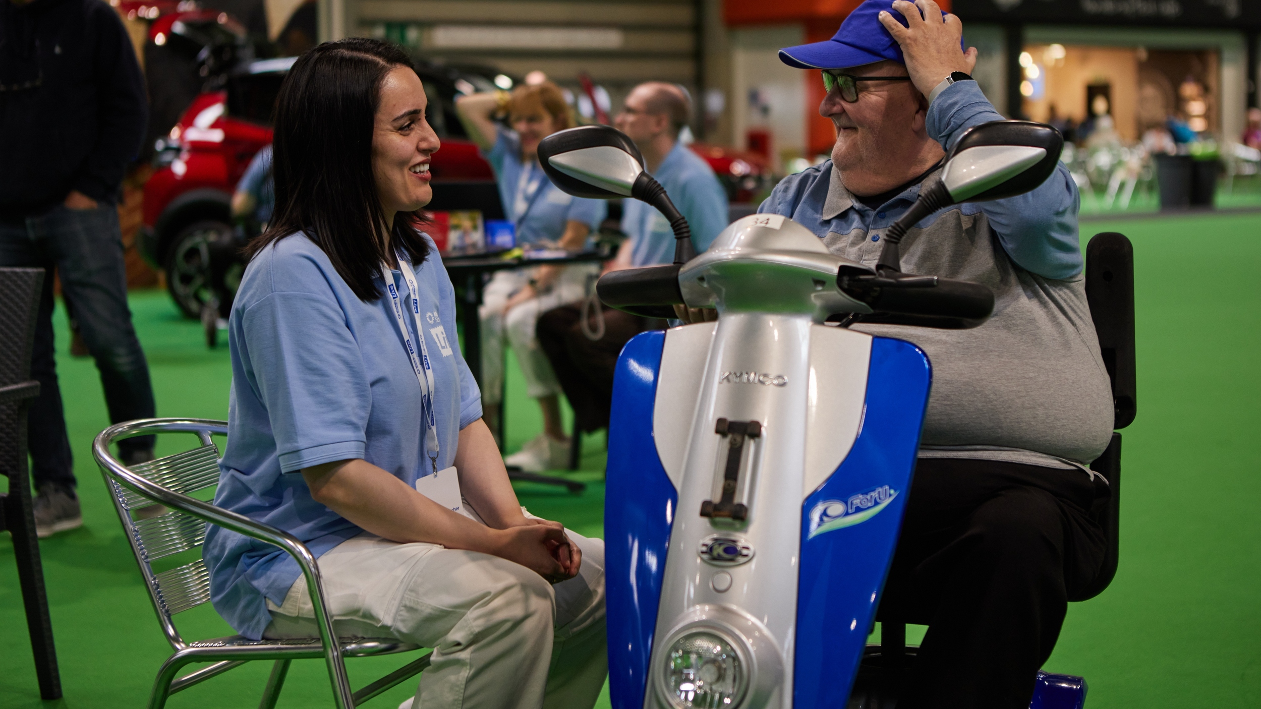 A man sits on a mobility scooter and smiles while talking to a staff member at Motability Scheme Live in Birmingham. Other visitors and vehicles are around them.