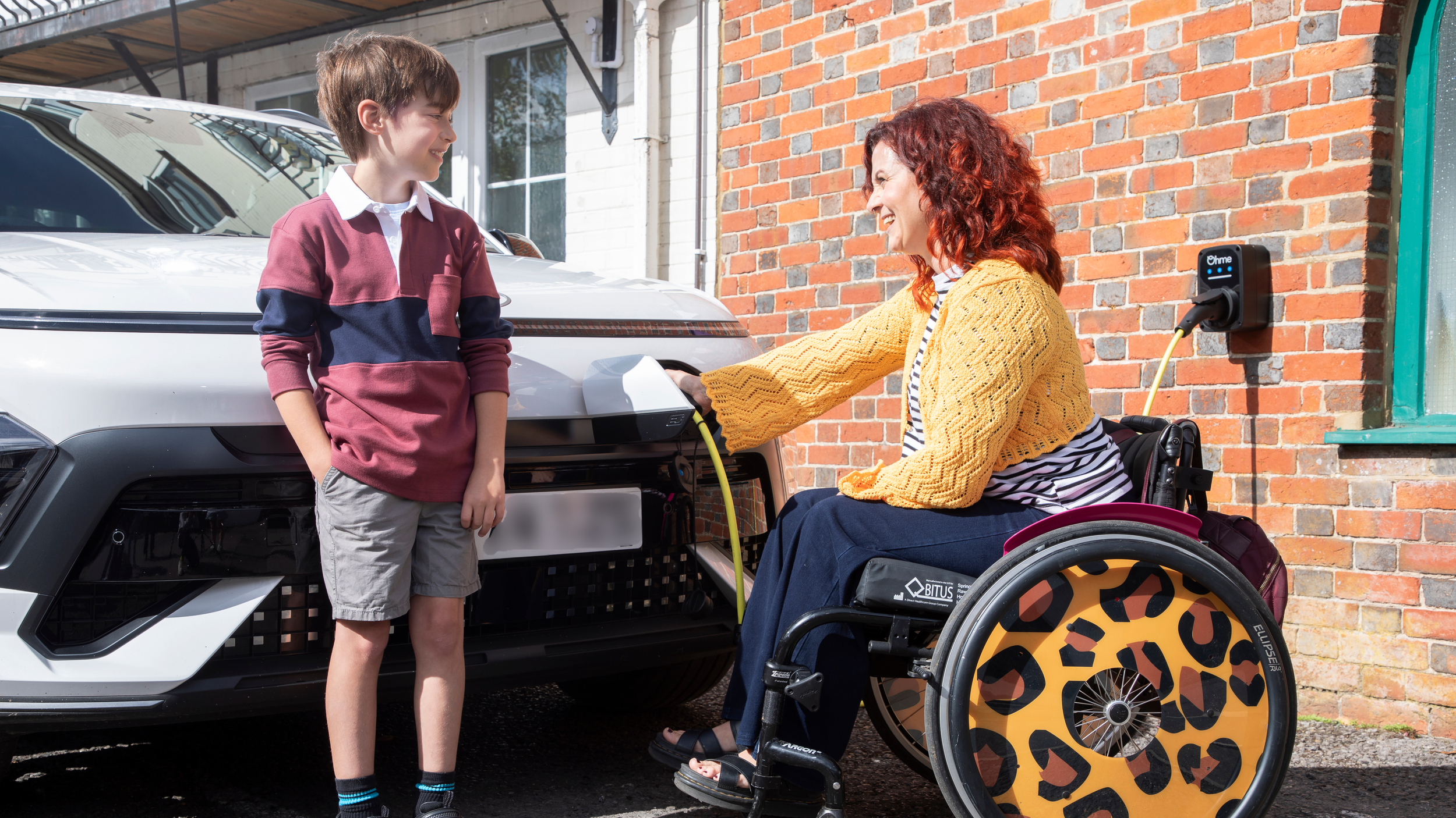 Woman with red curly hair sitting in a wheelchair connects a charging cable to a white electric car on a driveway, smiling at a young boy standing beside her. A home EV charger is mounted on the brick wall behind them, with the cable running from the wall unit to the car. The wheelchair has large wheels with a bold leopard print design.