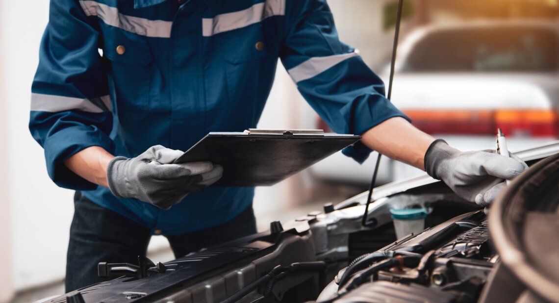Mechanic in a blue work uniform and gloves inspecting a car engine while holding a clipboard, with the bonnet open in a garage setting.