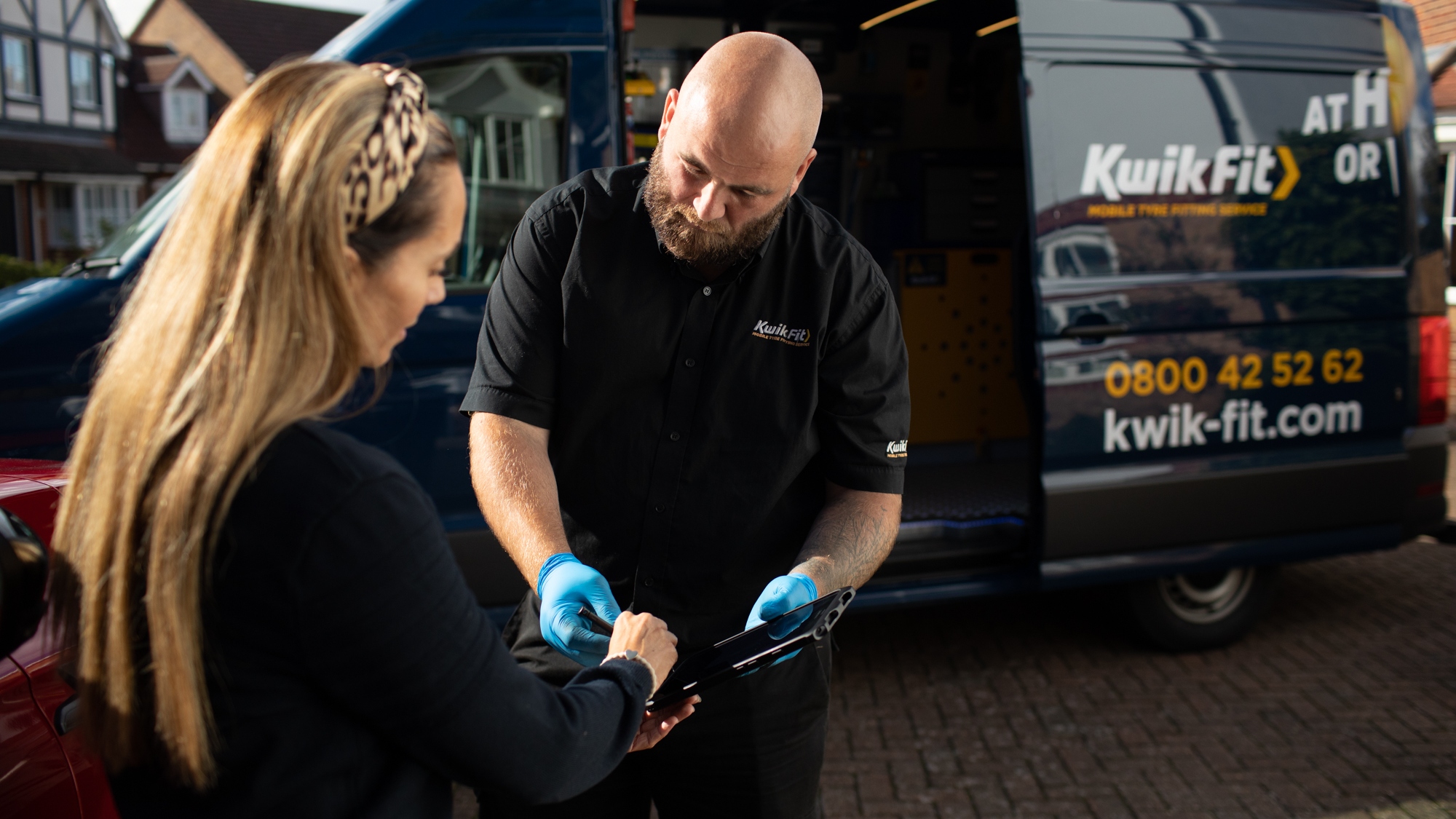 Kwik Fit mobile tyre technician wearing gloves checks a customer’s tyre and shows information on a tablet beside a branded service van parked on a residential driveway.