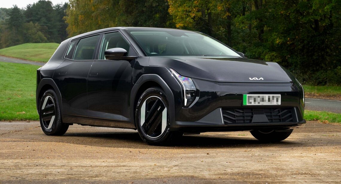 Black Kia EV4 electric car parked on a quiet rural road, shown from a front three quarter angle with a sleek modern design, distinctive LED lighting and aerodynamic wheels, surrounded by green trees in early autumn. Available on the Motability Scheme from £1,299 Advance Payment