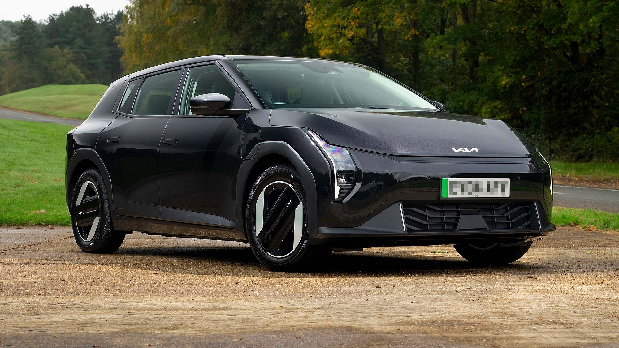 Black Kia EV4 electric car parked on a quiet rural road, shown from a front three quarter angle with a sleek modern design, distinctive LED lighting and aerodynamic wheels, surrounded by green trees in early autumn. Available on the Motability Scheme from £1,299 Advance Payment