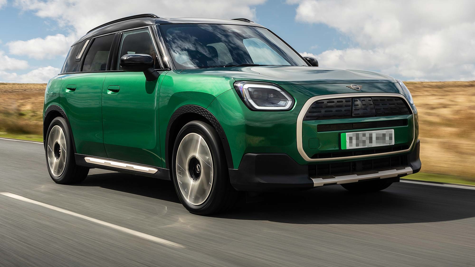 Green MINI Countryman SUV driving along a winding rural road with hills in the background, available on the Motability Scheme with no Advance Payment.