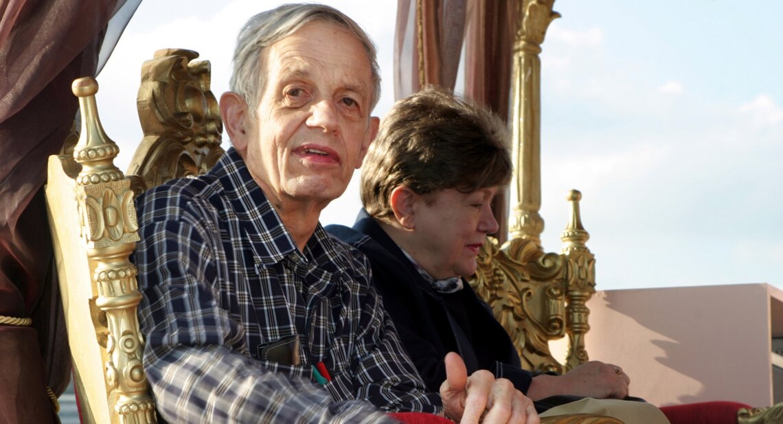 John Nash sits on an ornate gold chair wearing a checked shirt, looking toward the camera, with another person seated beside him facing away.