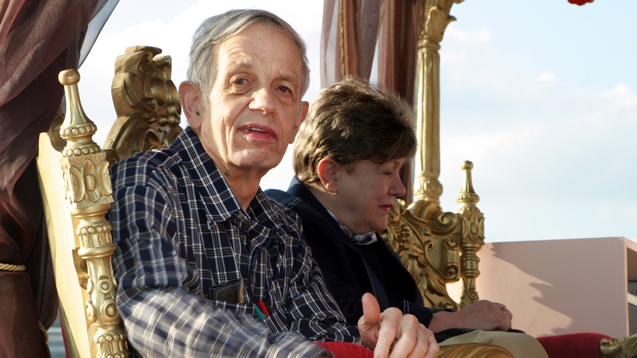 John Nash sits on an ornate gold chair wearing a checked shirt, looking toward the camera, with another person seated beside him facing away.