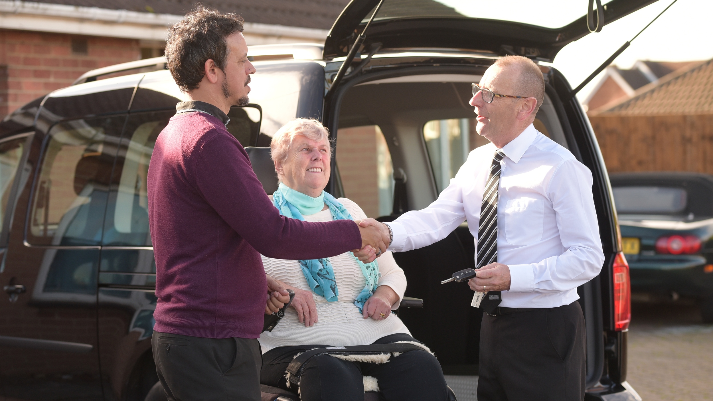 A Motability Scheme customer in a wheelchair smiles while seated at the rear of a black Wheelchair Accessible Vehicle (WAV) with its tailgate open, as a man shakes hands with a representative holding car keys during a home vehicle handover on a residential driveway.
