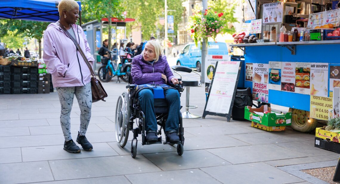 A woman using a powered wheelchair moves along a busy outdoor market street, turning to speak with a companion walking beside her. They pass a colourful juice and snack stall displaying fruit, smoothies and iced coffee, with other shoppers and market stands in the background.