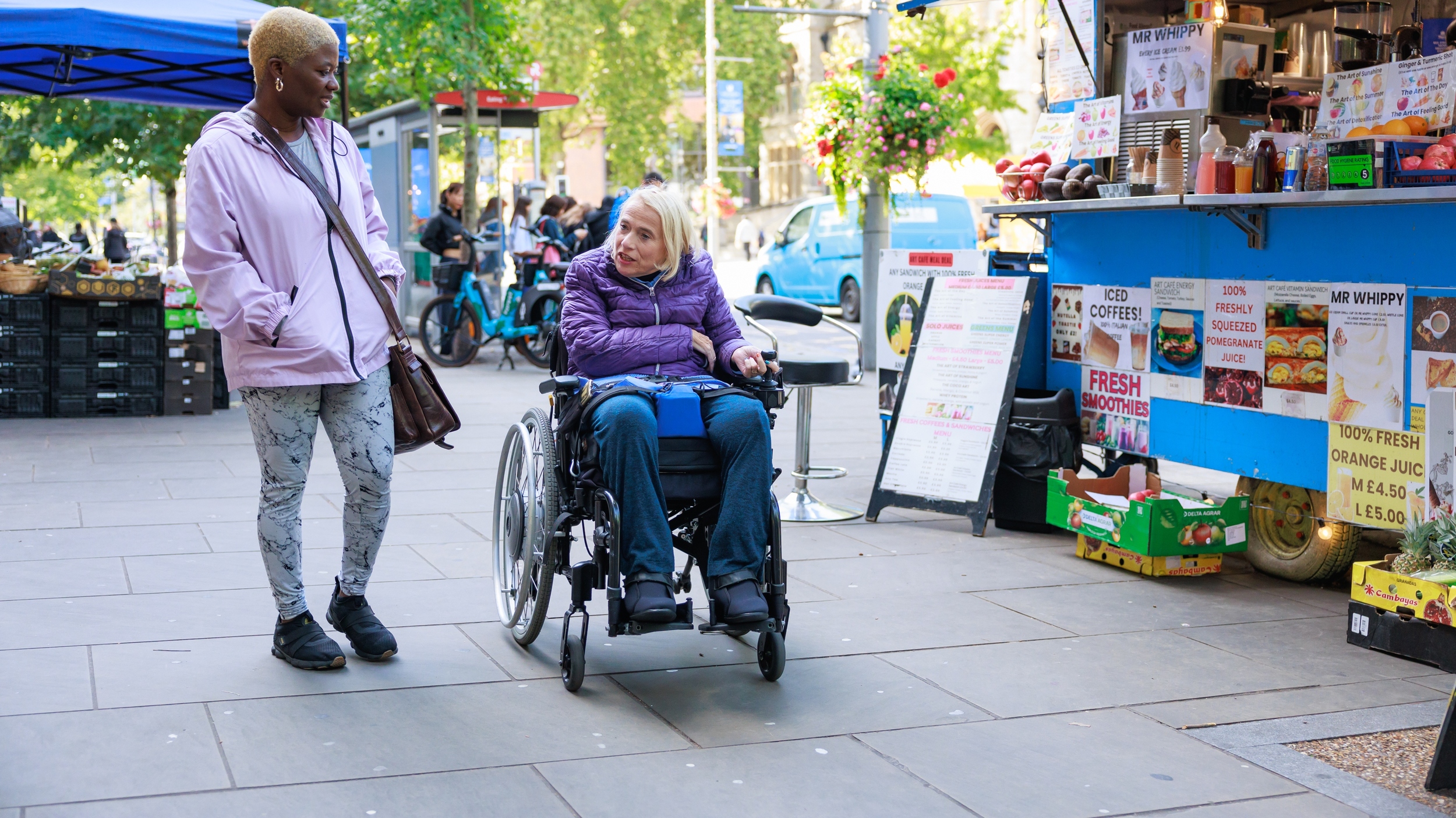 A woman using a powered wheelchair moves along a busy outdoor market street, turning to speak with a companion walking beside her. They pass a colourful juice and snack stall displaying fruit, smoothies and iced coffee, with other shoppers and market stands in the background.
