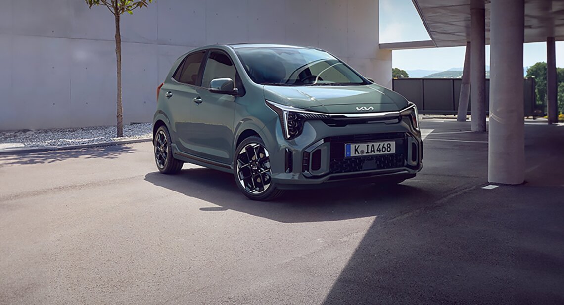 Green Kia Picanto parked outside a modern building with clean architecture, a small tree and shaded driveway area.