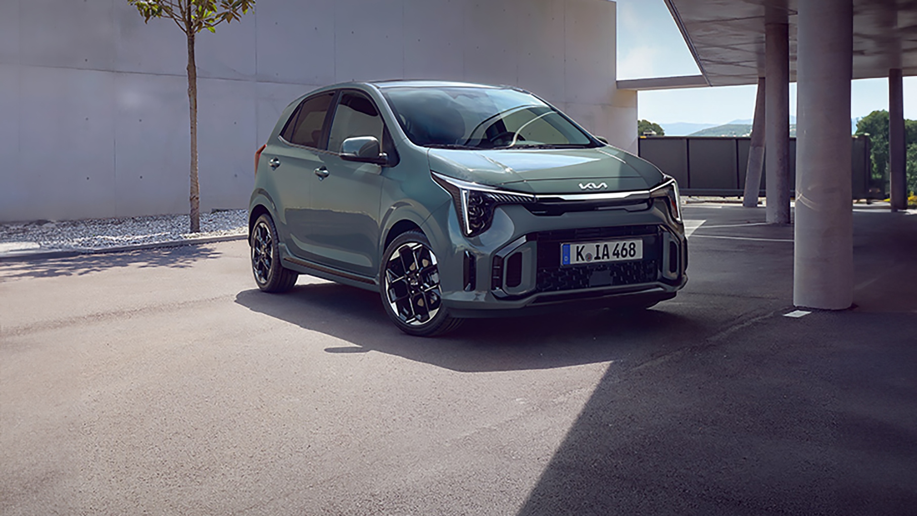 Green Kia Picanto parked outside a modern building with clean architecture, a small tree and shaded driveway area.