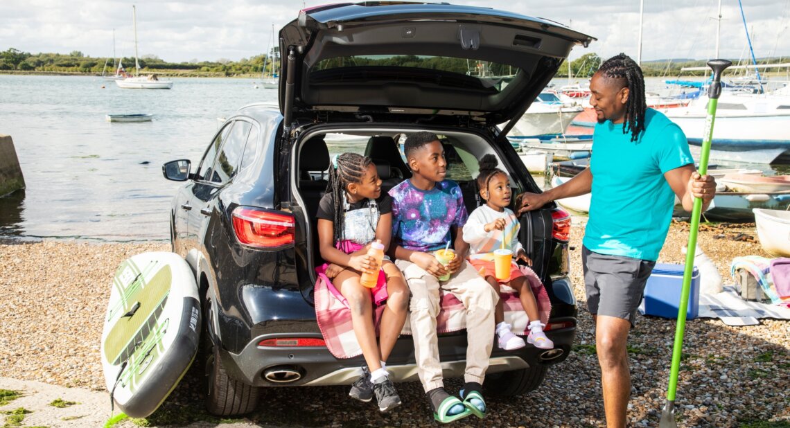 A father stands beside an open Motability Scheme car boot on a pebble beach, holding a paddle and smiling at three children sitting in the back with drinks. Paddleboards, a cooler box and boats on the water suggest a family day out by the shore.