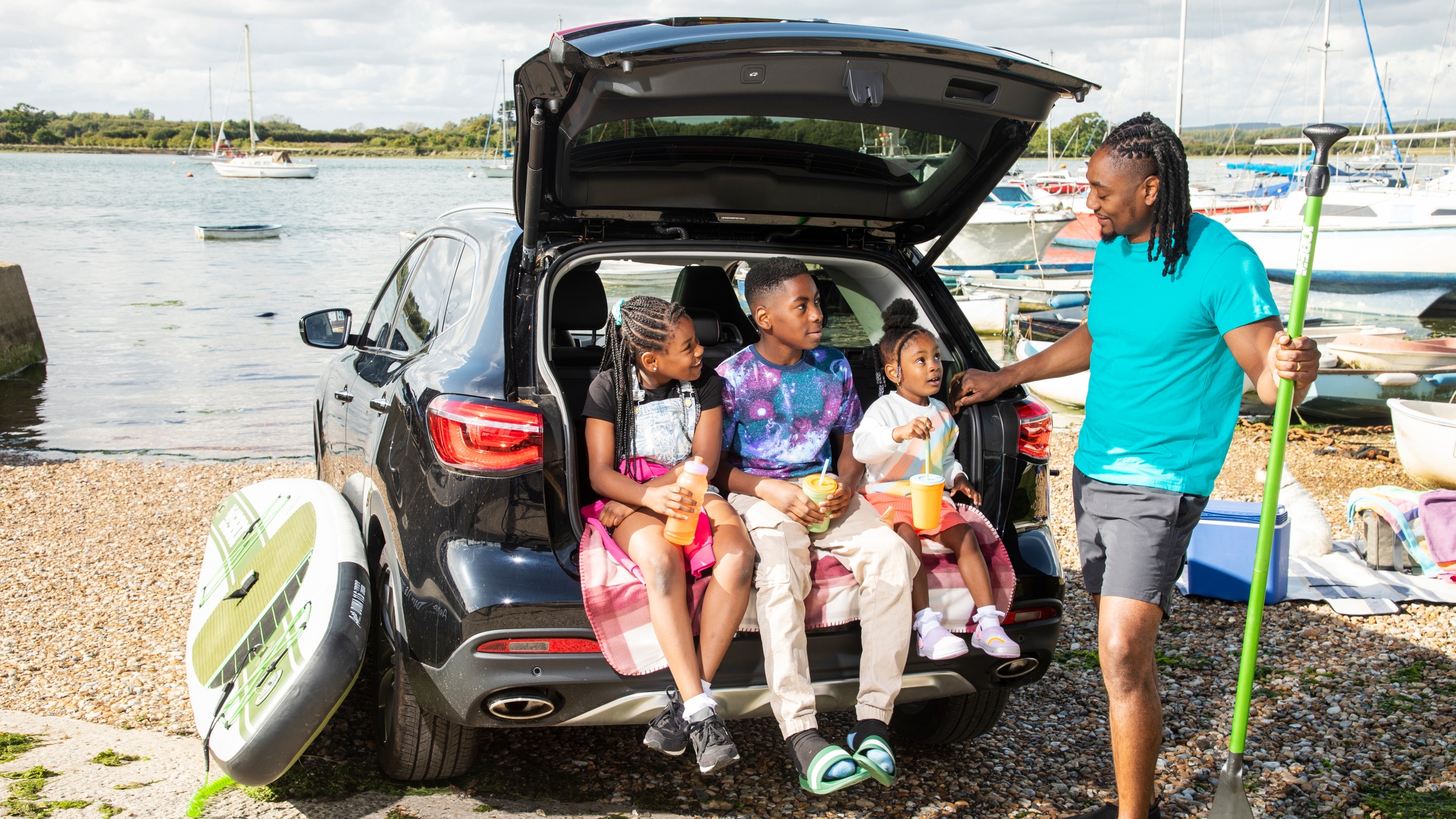 A father stands beside an open Motability Scheme car boot on a pebble beach, holding a paddle and smiling at three children sitting in the back with drinks. Paddleboards, a cooler box and boats on the water suggest a family day out by the shore.