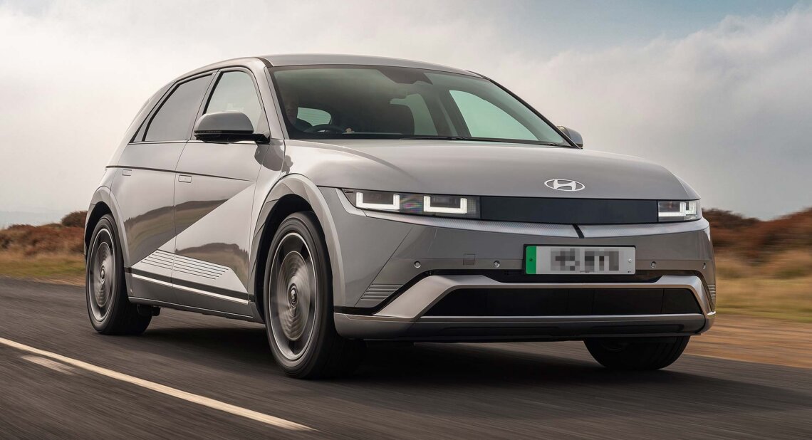 Grey Hyundai IONIQ 5 electric car driving on an open road, with countryside scenery and a cloudy sky behind. Available on the Motability Scheme from £3,499