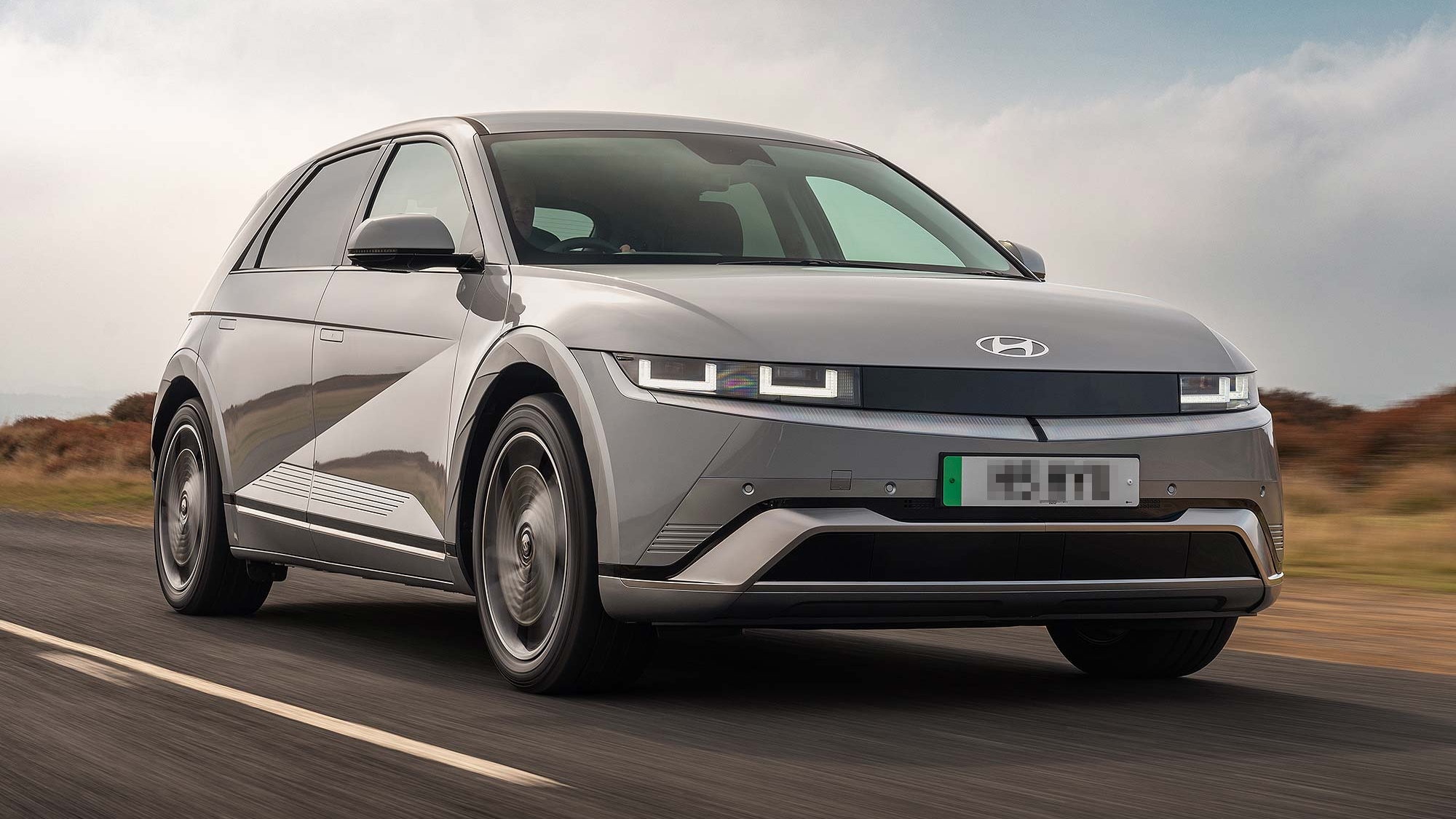 Grey Hyundai IONIQ 5 electric car driving on an open road, with countryside scenery and a cloudy sky behind. Available on the Motability Scheme from £3,499
