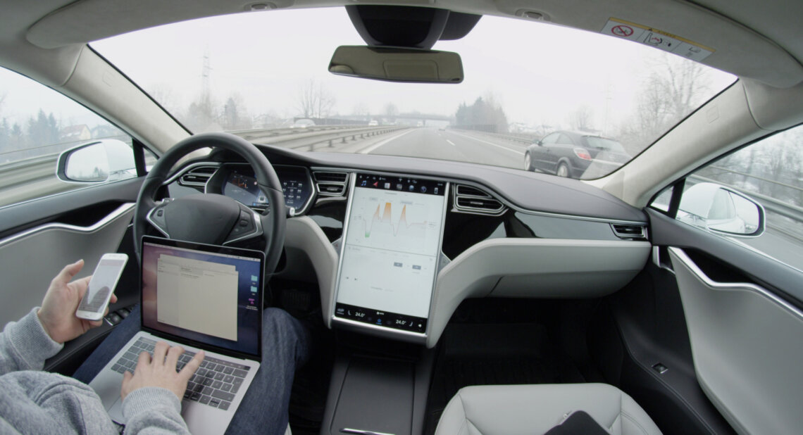 Driver using a laptop and phone inside a car on a motorway, with the steering wheel and dashboard visible.