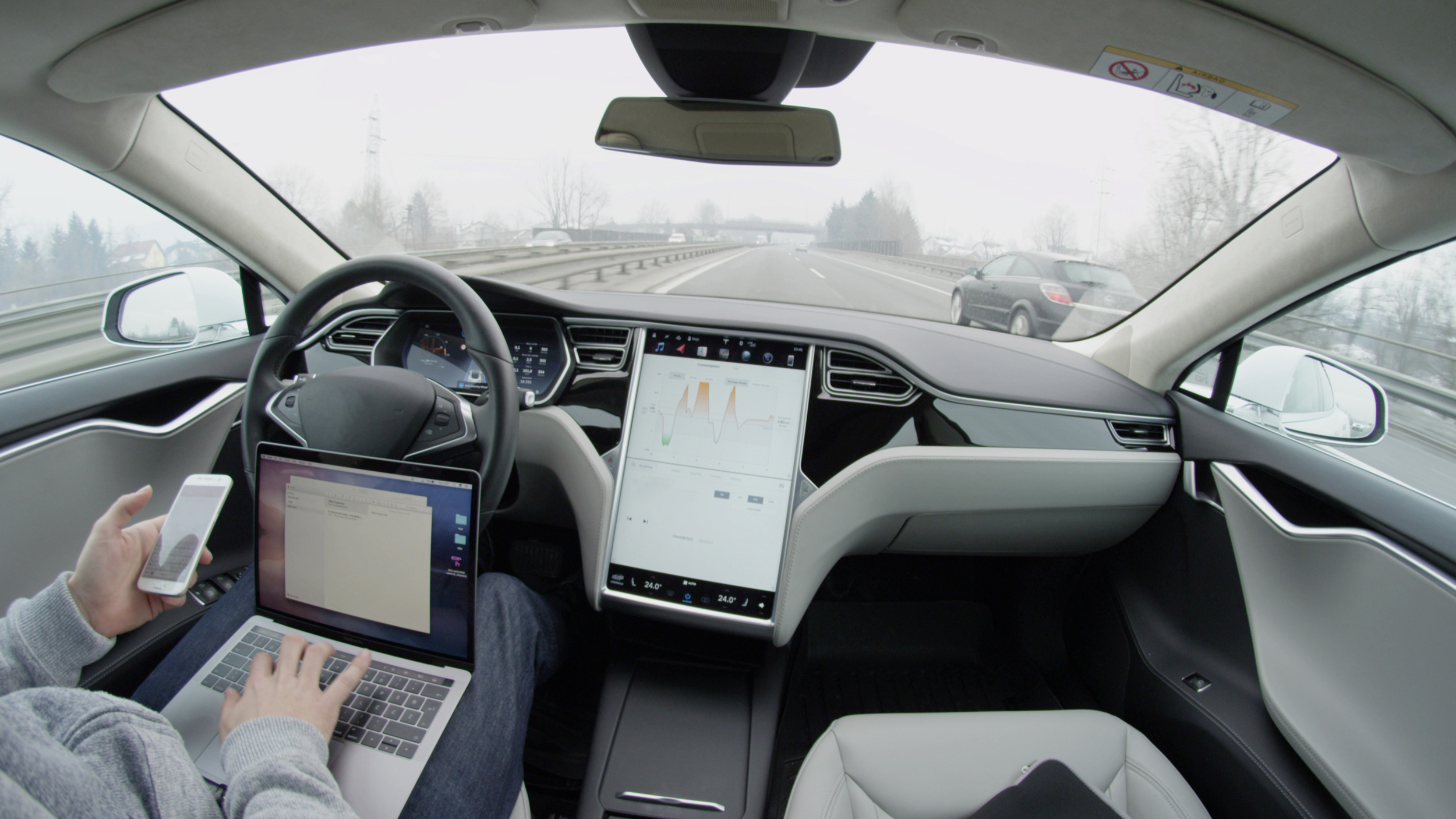 Driver using a laptop and phone inside a car on a motorway, with the steering wheel and dashboard visible.