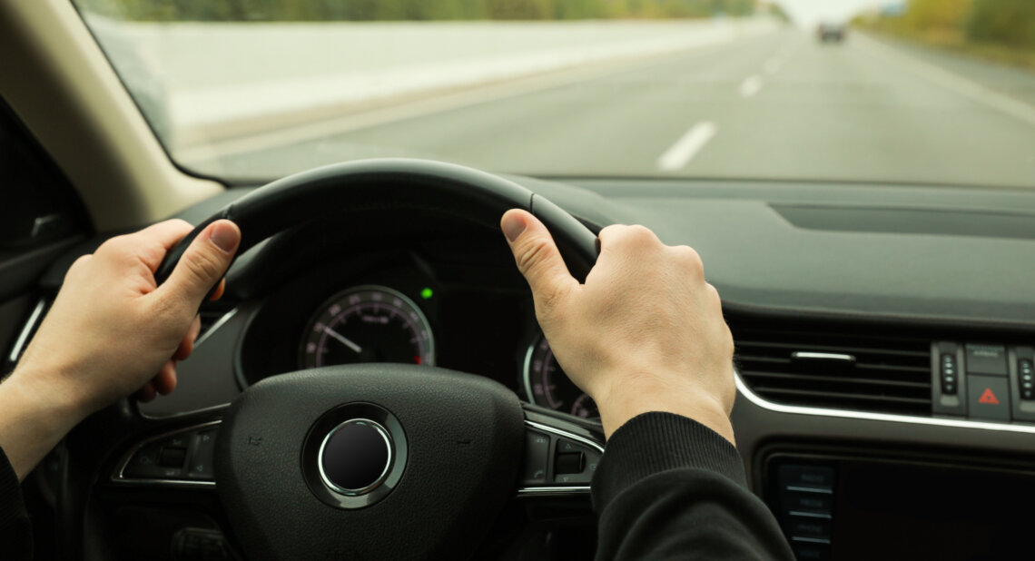 Driver holding a steering wheel on a quiet road, viewed from inside the car.