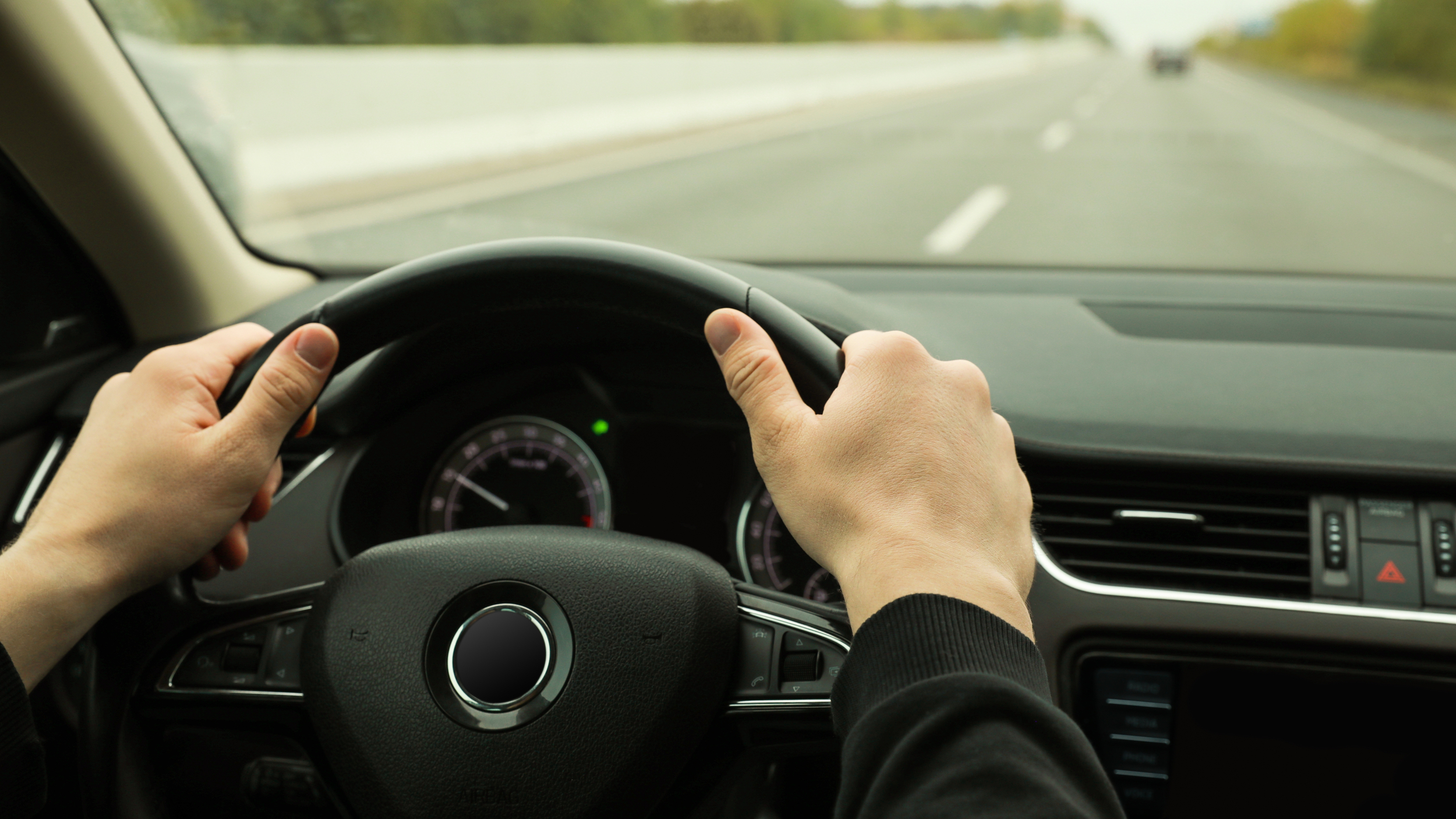Driver holding a steering wheel on a quiet road, viewed from inside the car.