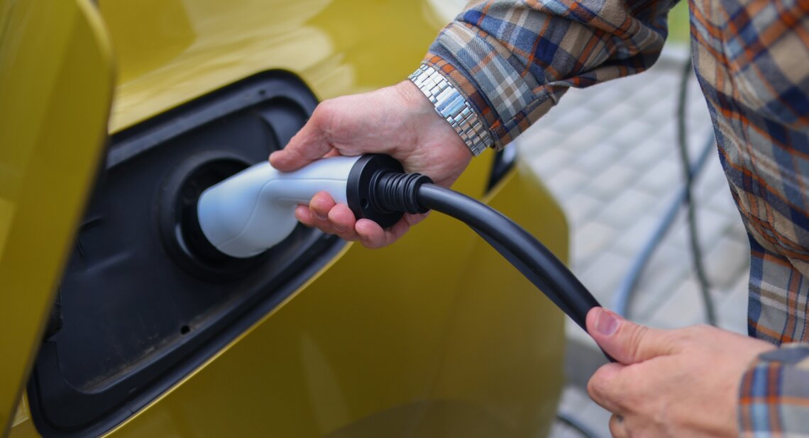 Close-up of a person plugging an electric vehicle charging cable into a yellow car, with their hands visible holding the connector.
