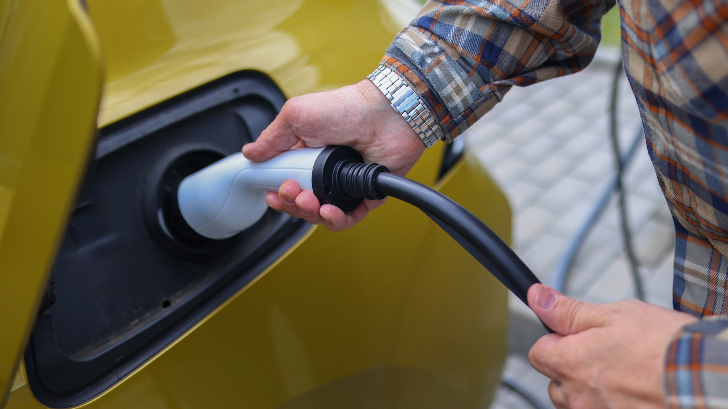 Close-up of a person plugging an electric vehicle charging cable into a yellow car, with their hands visible holding the connector.