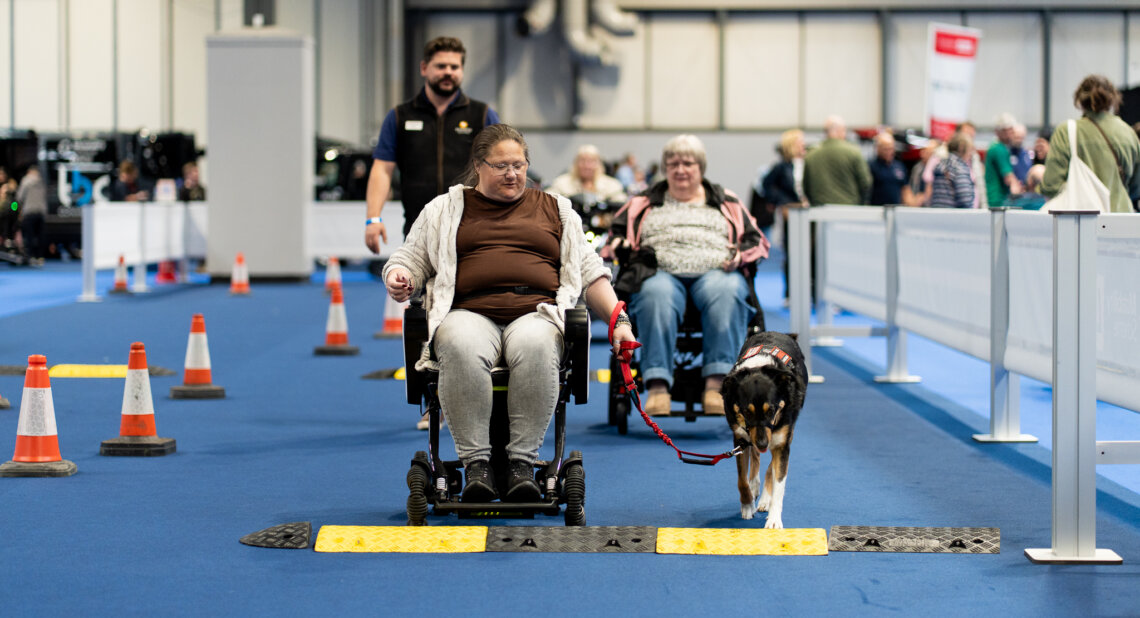 A person uses a powered wheelchair to drive over low obstacle ramps while a trained assistance dog walks beside them on a lead. Another wheelchair user follows behind with an instructor. Orange traffic cones mark the course inside a busy indoor exhibition hall.