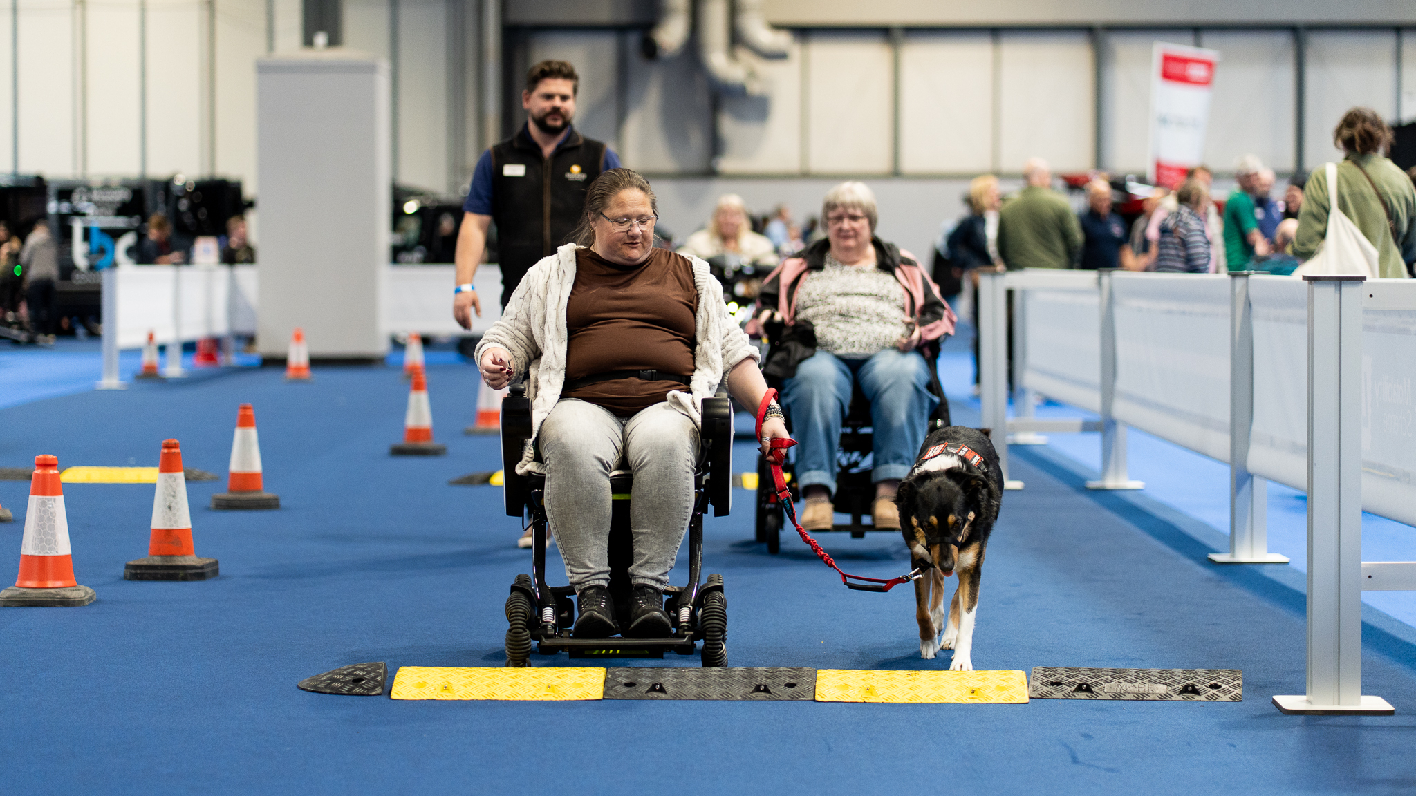 A person uses a powered wheelchair to drive over low obstacle ramps while a trained assistance dog walks beside them on a lead. Another wheelchair user follows behind with an instructor. Orange traffic cones mark the course inside a busy indoor exhibition hall.