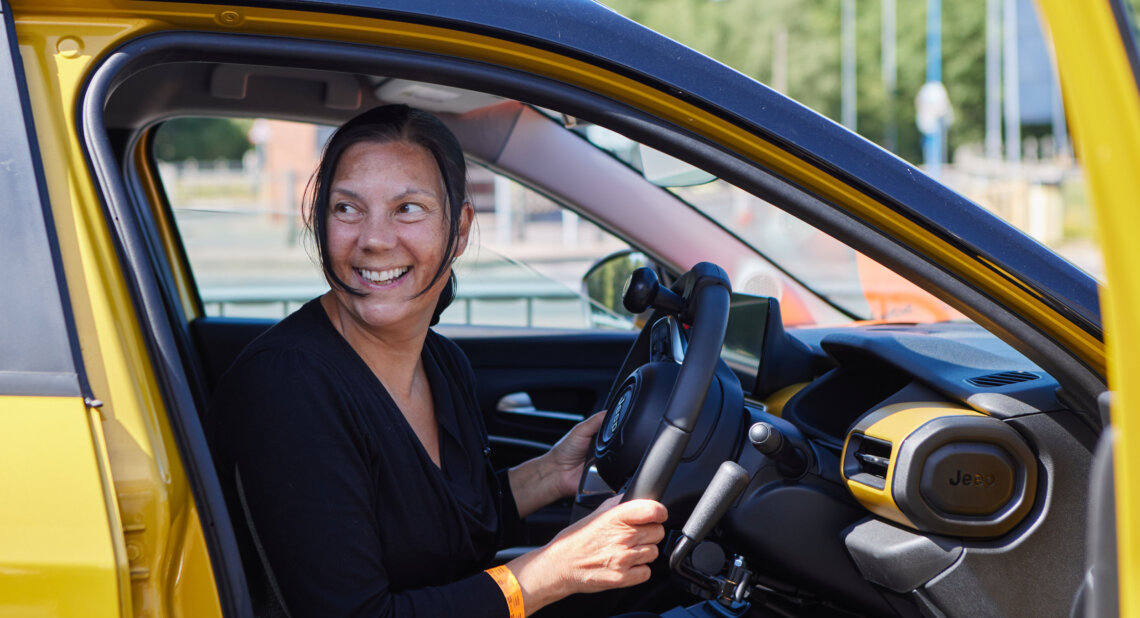 A smiling woman sitting in the driver’s seat of a yellow car. She is holding the steering wheel and looking out through the open door.