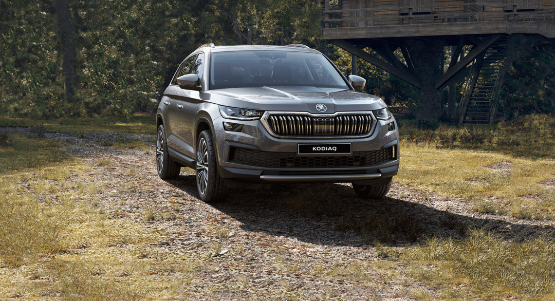 Silver Škoda Kodiaq SUV parked on a woodland track beside a large wooden treehouse structure. The vehicle is shown from the front, surrounded by trees and natural scenery.