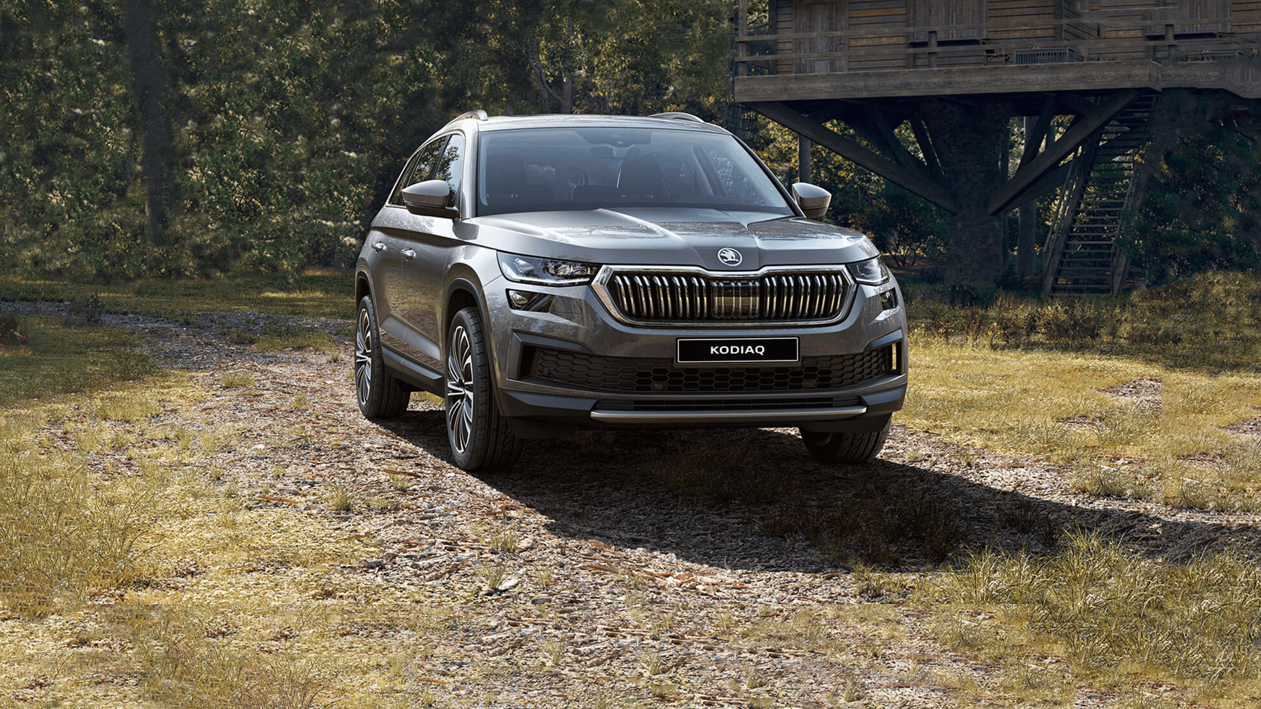 Silver Škoda Kodiaq SUV parked on a woodland track beside a large wooden treehouse structure. The vehicle is shown from the front, surrounded by trees and natural scenery.