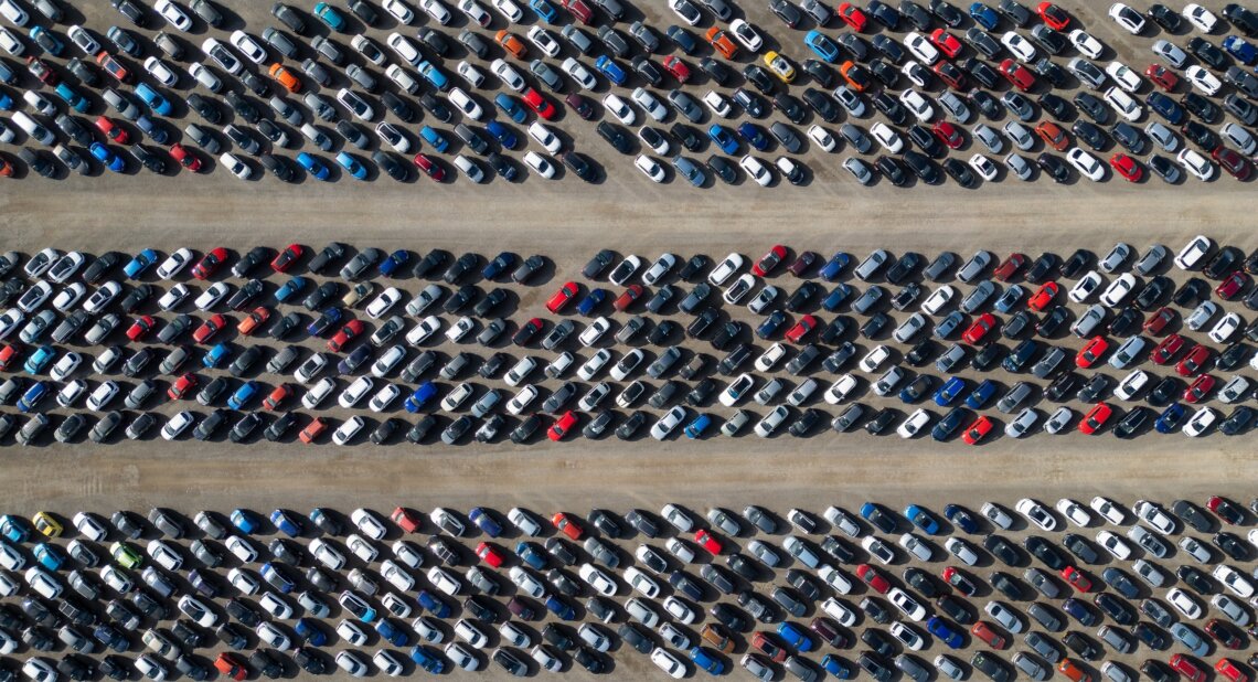 Aerial view of a large outdoor car park with hundreds of cars parked in long, diagonal rows separated by wide access lanes. Most vehicles are white, black, grey or blue, with a few red, orange and yellow cars adding contrast. The image fills the frame with repeating patterns of tightly parked vehicles, viewed from directly above.